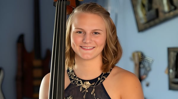 Eine junge Frau hält eine Violine vor einer Wand mit gerahmten Kunstwerken im Queensbury Union Free School District.