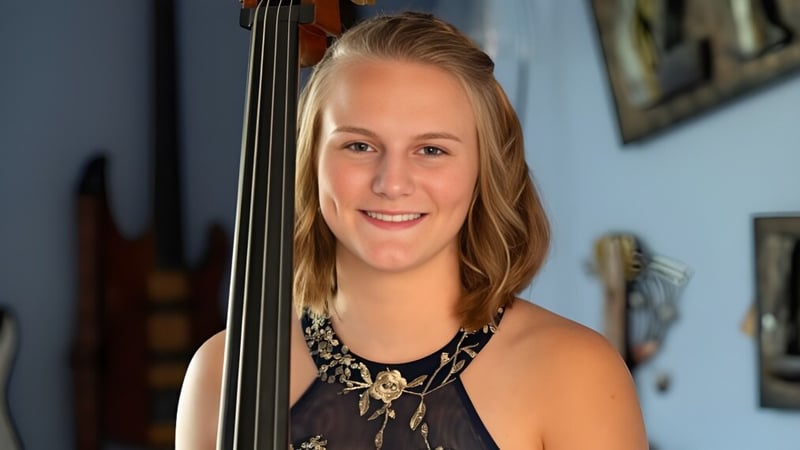 Eine junge Frau hält eine Violine vor einer Wand mit gerahmten Kunstwerken im Queensbury Union Free School District.