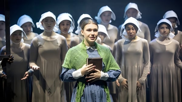 Eine Frau mit gruenem Schal haelt ein Buch vor einer Gruppe von Frauen in weissen Gewaendern auf dem Campus der Queenswood School.