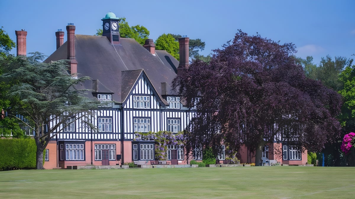 Das Tudor-Gebäude der Queenswood School steht umgeben von grünen Bäumen und einem Baum mit lila Blättern unter blauem Himmel.