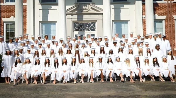 Schüler der Rabun Gap-Nacoochee School in weißen Abschlusskleidern versammeln sich vor einem historischen Backsteingebäude.