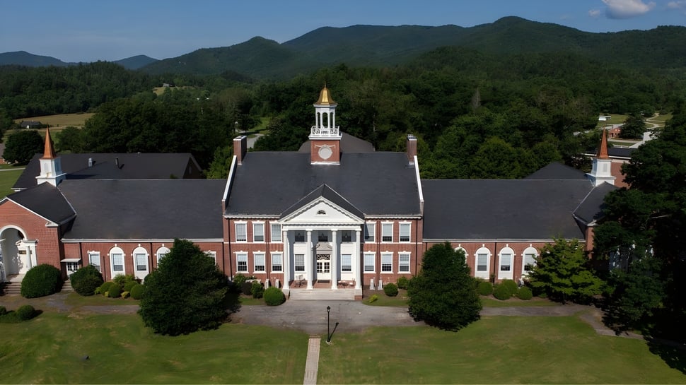 Das Hauptgebäude der Rabun Gap-Nacoochee School steht in einer grünen Landschaft mit Bergen im Hintergrund.