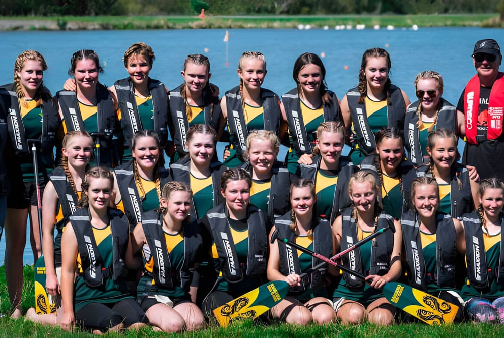 Eine Gruppe junger Sportlerinnen in grünen und gelben Trikots posiert vor einem See mit Bäumen auf dem Gelände der Rangiora High School.