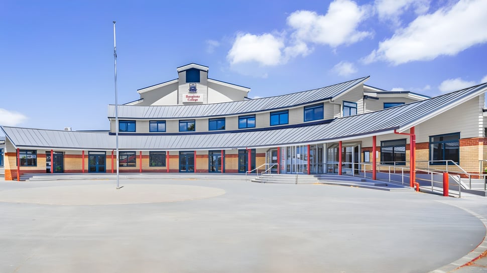 Das moderne mehrstöckige Gebäude mit geschwungenem Dach und großen Fenstern des Rangitoto College ist von einem gepflasterten Außenbereich umgeben.