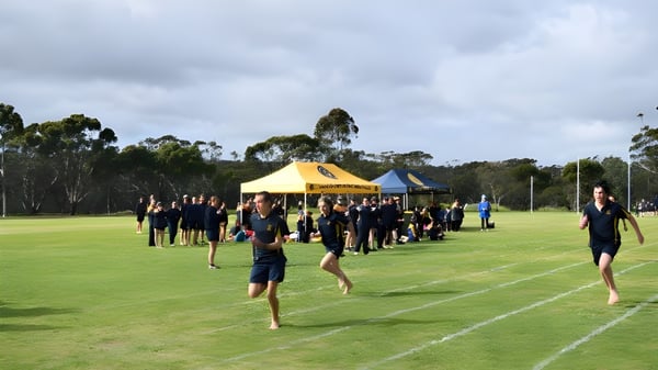 Schülerinnen und Schüler der Ravensthorpe District High School versammeln sich in Sportkleidung auf einem Rasenfeld mit Zelten und Bäumen im Hintergrund.