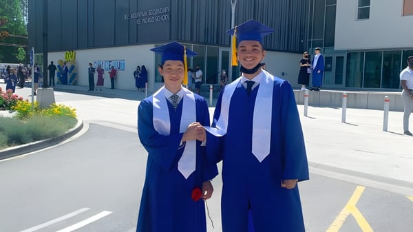Zwei Schüler in Abschlussroben stehen vor dem Gebäude der R.E. Mountain Secondary School.