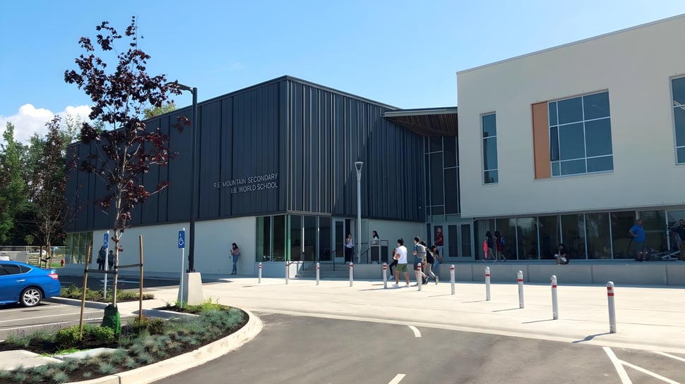 Das moderne mehrstöckige Gebäude der R.E. Mountain Secondary School mit dunkler Fassade und umgebender Pflasterfläche ist zu sehen.
