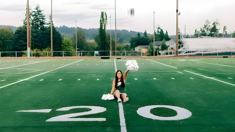 Eine Cheerleaderin der Redmond High School steht auf dem Kunstrasenfeld vor einem Waldstück und Gebäuden.