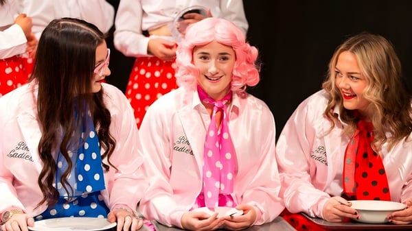 Drei Schülerinnen der Reed’s School tragen bunte gepunktete Outfits und stehen lächelnd auf einer Bühne mit pink-weißem Hintergrund.