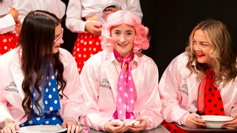 Drei Schülerinnen der Reed’s School tragen bunte gepunktete Outfits und stehen lächelnd auf einer Bühne mit pink-weißem Hintergrund.