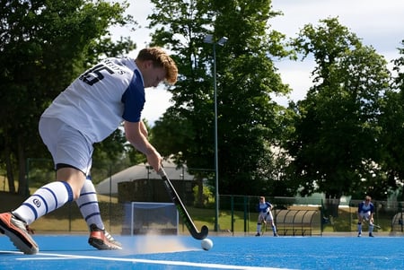 Ein Schüler der Reed’s School spielt Feldhockey auf einem blauen Kunstrasenplatz mit anderen Spielern im Hintergrund.