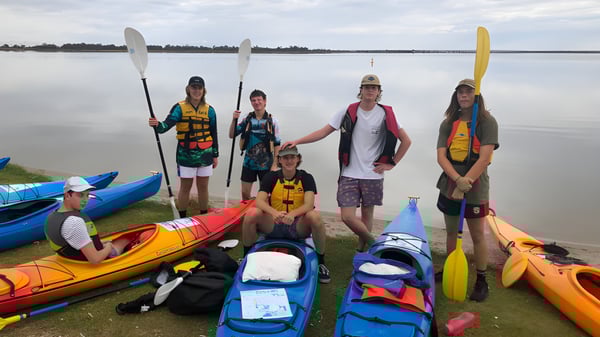 Schüler der Renmark High School stehen am Seeufer zwischen bunten Kajaks und Paddleboards in einer ruhigen Landschaft.