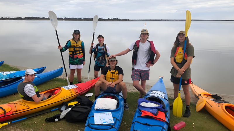 Schüler der Renmark High School stehen am Seeufer zwischen bunten Kajaks und Paddleboards in einer ruhigen Landschaft.