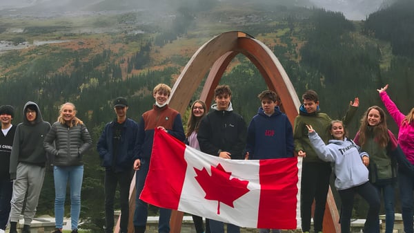 Schüler der Revelstoke Secondary School stehen vor einem großen Holzbogen und halten eine kanadische Flagge vor bergigem Waldlandschaft.