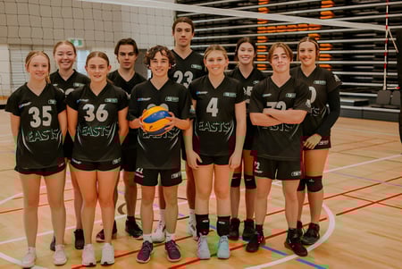 Eine Gruppe junger Volleyballspielerinnen des Reynella East College steht gemeinsam auf dem Volleyballfeld mit Netz und Anzeigetafel im Hintergrund.
