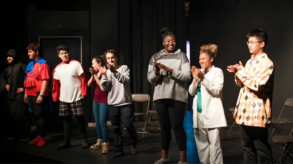 Schüler der Ribet Academy stehen auf einer Bühne in einem schwach beleuchteten Raum mit Vorhängen und Bildern im Hintergrund.