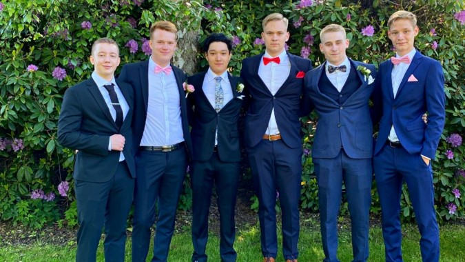 Eine Gruppe junger Männer im formellen Anzug steht in den Gartenanlagen des Rice College.