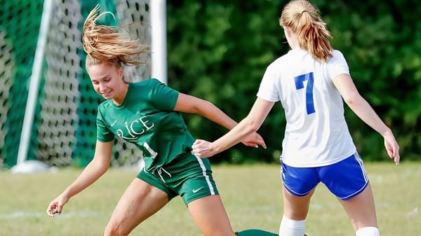 Zwei Schülerinnen der Rice Memorial High School spielen Fußball auf einem Feld mit sichtbarem Tor im Hintergrund.