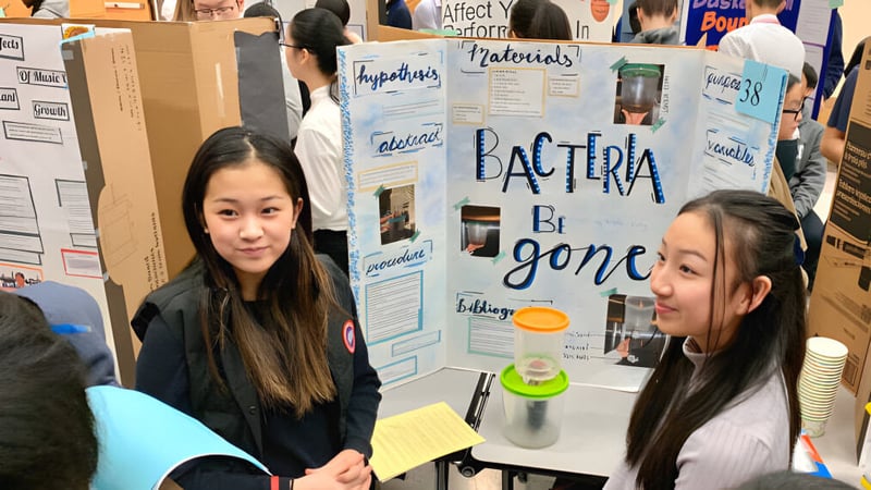 Zwei Schülerinnen der Richmond Secondary School stehen vor einem Informationstafel über Bakterien mit wissenschaftlichen Materialien.