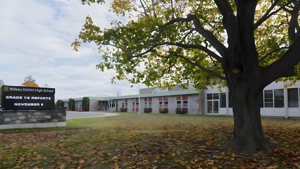 Ein großer Baum mit herbstlich gefärbten Blättern steht vor dem Schulgebäude der Rideau District High School mit einem Berichtshinweis für Jahrgang 7-8.