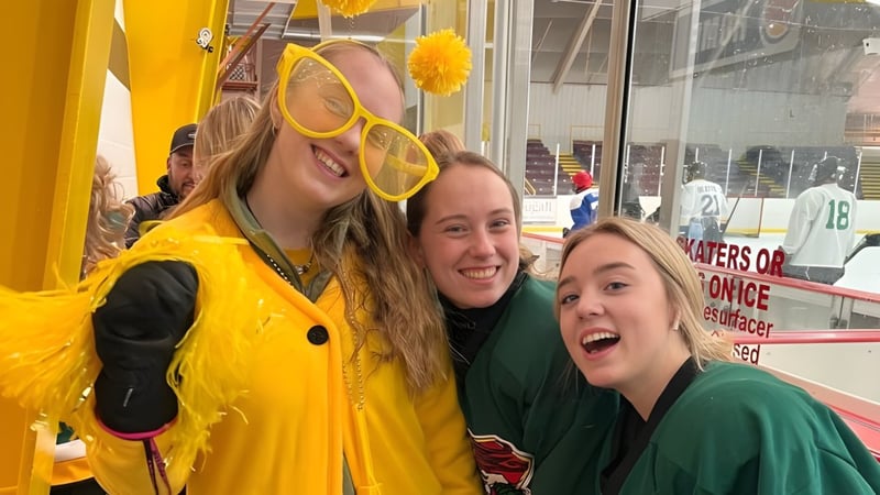 Drei Schülerinnen der Rideau District High School posieren lächelnd in einer Sportarena mit Sportgeräten im Hintergrund.