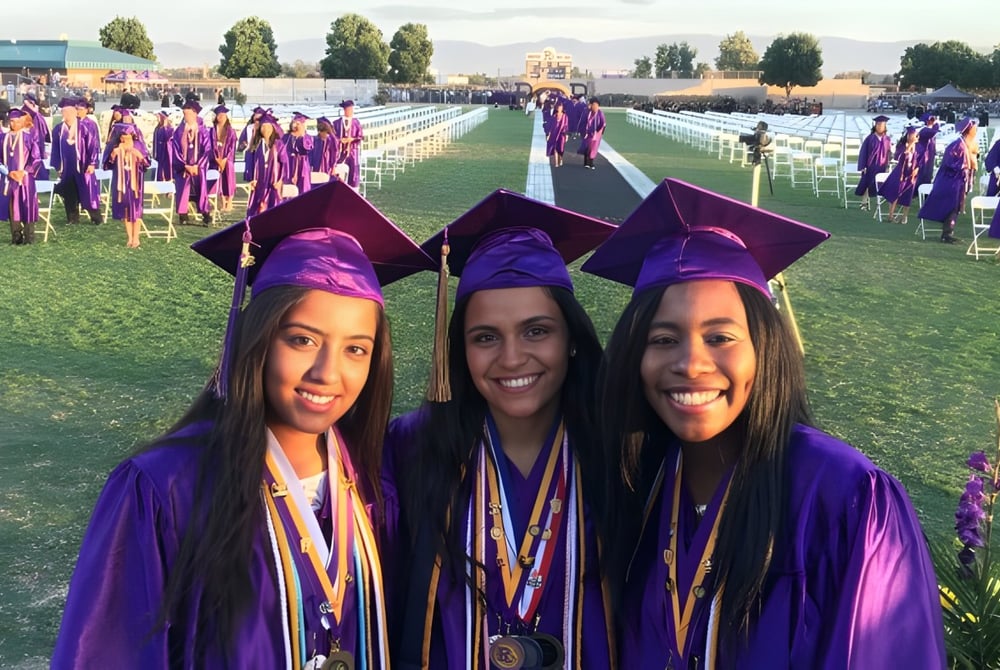 Drei Absolventinnen der Ridgeview High School in lila Abschlusskleidung stehen vor einer großen Gruppe von Absolventen auf einem Grasfeld.