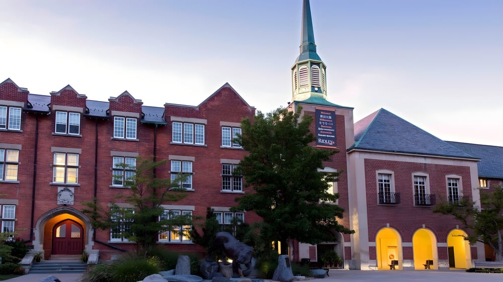 Das historische Backsteingebäude mit hohem Turm auf dem Gelände des Ridley College ist von Bäumen und weiteren Gebäuden umgeben.