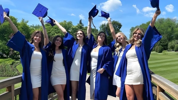Eine Gruppe von Absolventinnen der River East Collegiate feiert im Freien mit erhobenen Armen und Diplomen.