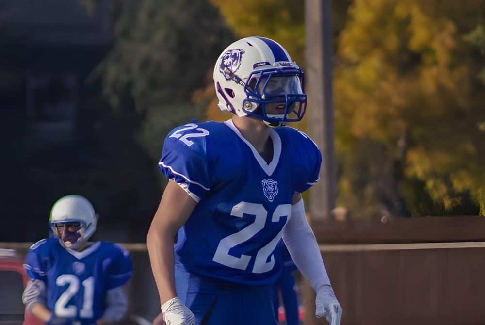 Ein Footballspieler in blauem Trikot steht bei Nacht auf dem Spielfeld der River East Collegiate mit anderen Spielern im Hintergrund.