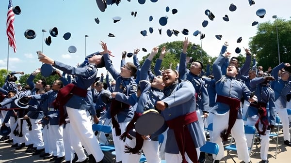 Absolventinnen und Absolventen der Riverside Military Academy werfen ihre Abschlussmützen in die Luft vor einer amerikanischen Flagge.