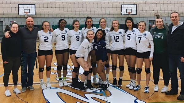 Schülerinnen der Riverside Secondary School stehen in ihren Volleyball-Uniformen auf einem Basketballfeld mit Körben im Hintergrund.
