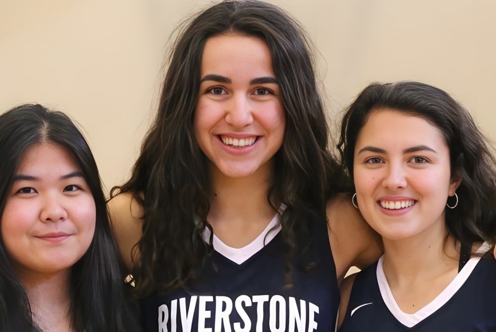 Drei Schülerinnen mit dunklem Haar lächeln und posieren zusammen in einer entspannten Umgebung auf dem Campus der Riverstone International School.