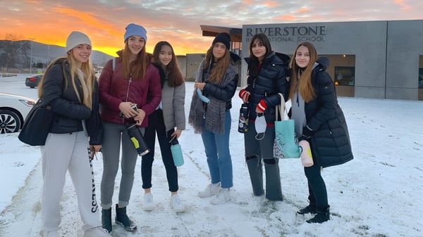 Eine Gruppe Schülerinnen steht draußen im Schnee vor der Riverstone International School mit einem bunten Sonnenuntergang im Hintergrund.
