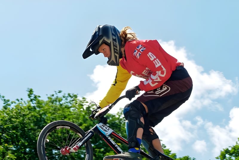 Eine Person in buntem Trikot macht einen Sprung mit dem Fahrrad vor grünem Laub und bewölktem Himmel auf dem Gelände von Rochester Independent College.