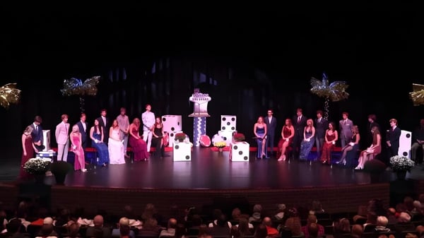 Schüler der Rockford High School treten in farbenfrohen Kostümen mit aufwendigen Kopfschmuck auf einer Bühne mit dekorativem Hintergrund auf.