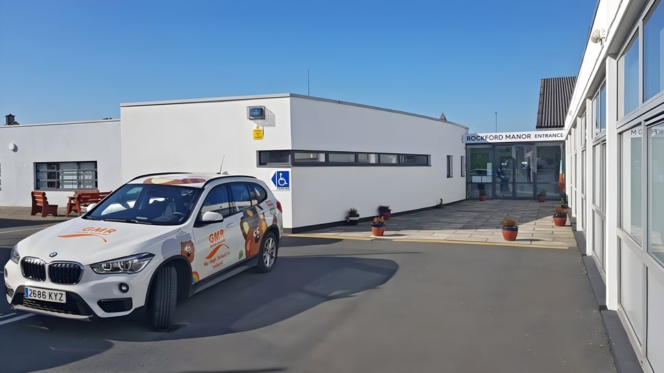 Ein weißer BMW X1 steht vor einem großen weißen Gebäude auf dem Campus der Rockford Manor School bei klarem Himmel.