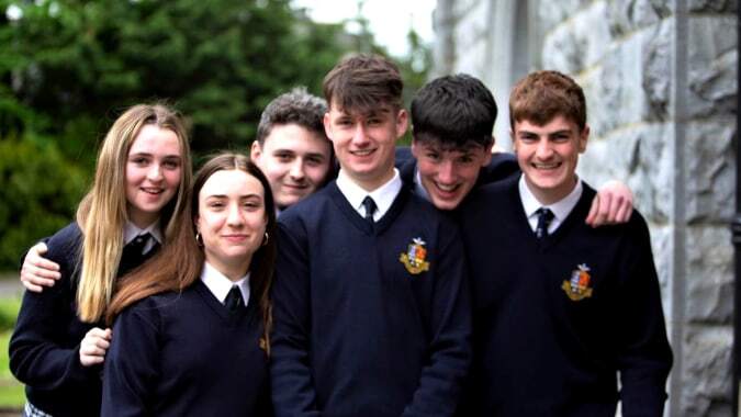 Eine Gruppe Schülerinnen und Schüler in Schuluniformen steht vor einer Steinmauer auf dem Campus des Rockwell College.