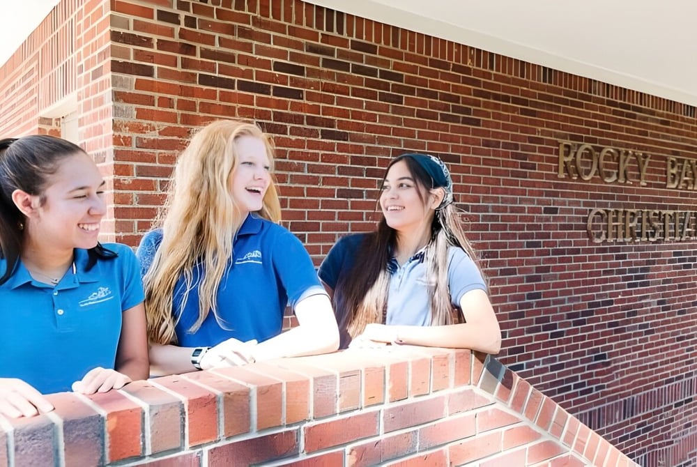 Drei Schülerinnen der Rocky Bayou Christian High School stehen gemeinsam vor einer Backsteinmauer.