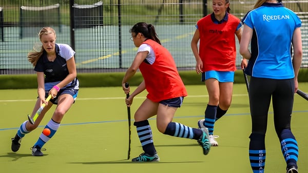 Schülerinnen der Roedean School spielen Feldhockey auf einem Kunstrasenplatz mit Zaun und Bäumen im Hintergrund.