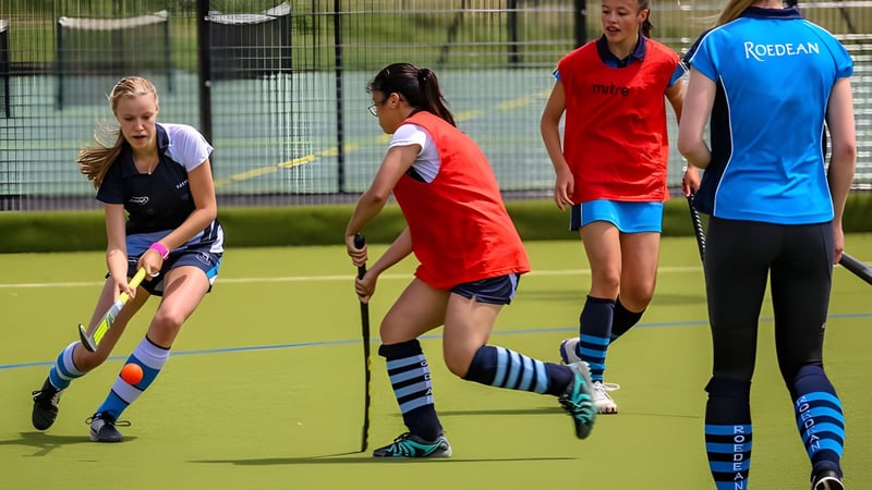Schülerinnen der Roedean School spielen Feldhockey auf einem Kunstrasenplatz mit Zaun und Bäumen im Hintergrund.