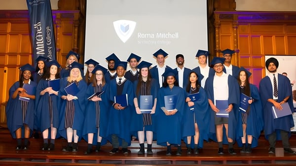 Absolventen in blauen Talaren stehen auf der Bühne vor einer Leinwand mit dem Logo der Roma Mitchell Secondary College.