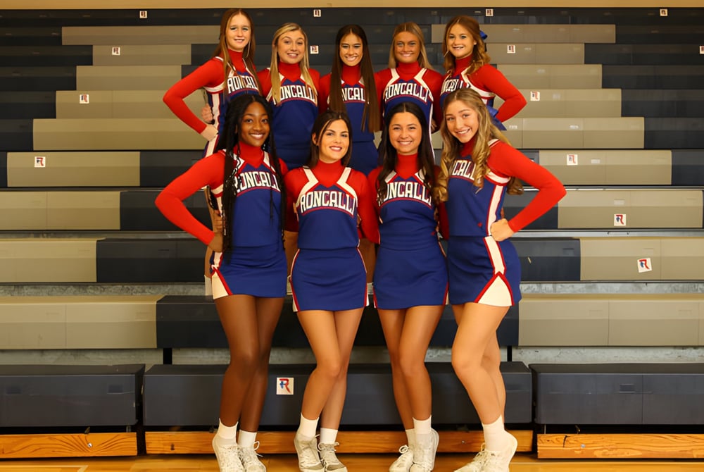 Schülerinnen der Roncalli Catholic High School stehen in roten und blauen Cheerleading-Uniformen auf einer Bühne vor einem dunklen Hintergrund.