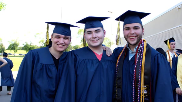Drei Schüler der Rosemount High School feiern ihren Abschluss in Talaren auf dem Schulgelände mit Grünflächen im Hintergrund.