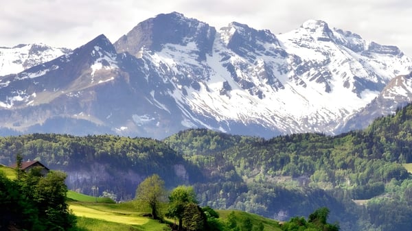 Eine grüne Landschaft mit Bäumen und einem kleinen Gebäude vor schneebedeckten Bergen auf dem Gelände der Ross Sheppard High School.
