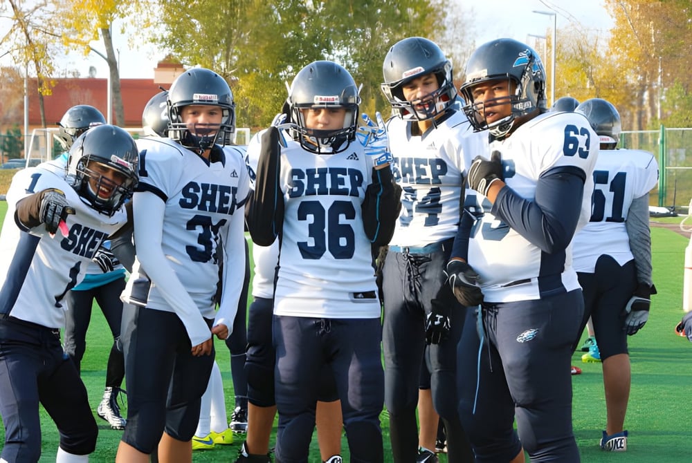 Eine Gruppe von Fußballspielern der Ross Sheppard High School steht gemeinsam auf einem Rasenfeld mit Bäumen und Gebäuden im Hintergrund.