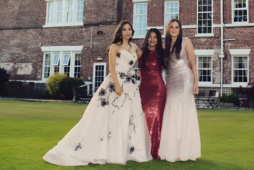 Drei Schülerinnen der Rossall School in festlichen Kleidern stehen vor einem Backsteingebäude mit großen Fenstern auf einer Rasenfläche.