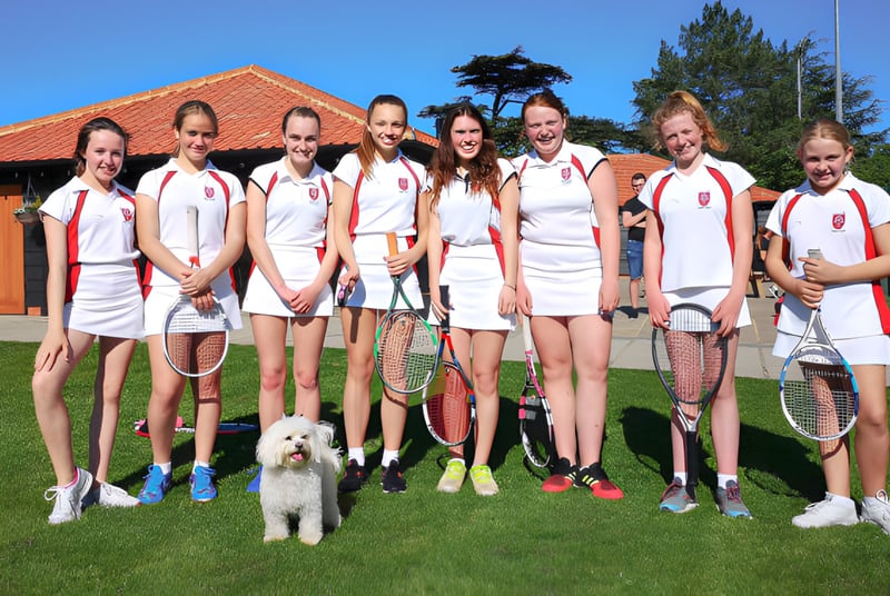 Eine Gruppe von Schülerinnen und Schülern der Royal Hospital School in Tennisbekleidung steht mit einem Hund auf einem Rasen vor einem Gebäude mit rotem Ziegeldach.