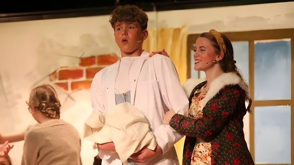 Zwei Personen stehen auf der Bühne der Royal School Armagh vor einer Backsteinwand mit Bühnenrequisiten im Hintergrund.