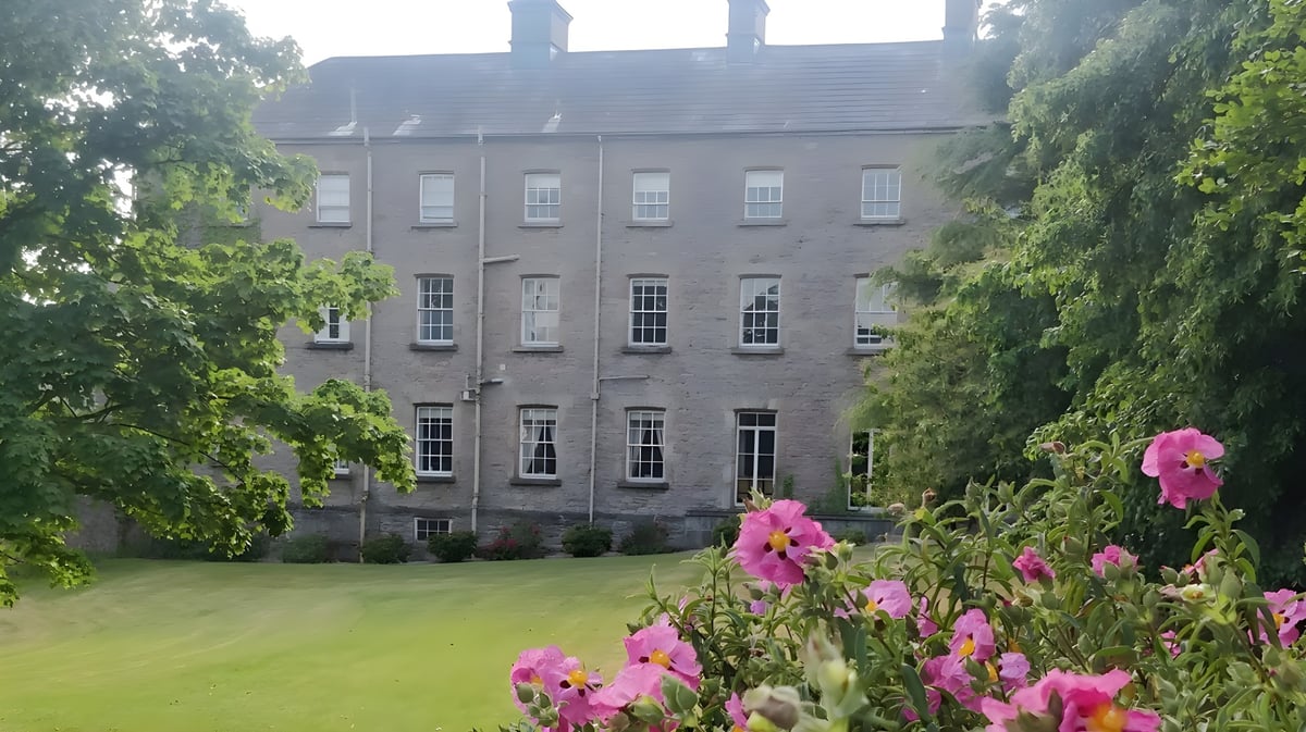 Das graue steinerne Gebäude mit vielen Fenstern steht auf dem Campus der Royal School Armagh umgeben von grünen Pflanzen und pinken Blumen.