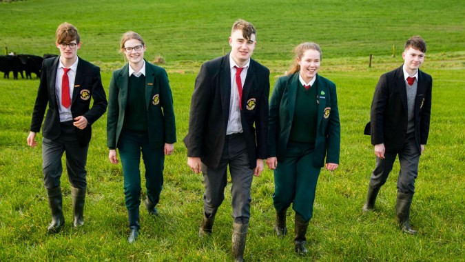 Schüler der Royal School Cavan stehen in Schuluniformen auf einer Wiese mit einer Herde Rinder im Hintergrund.
